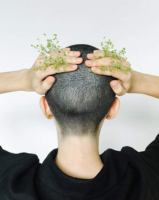Eine Person mit kurzgeschorenen Haaren legt die Handflächen auf den Hinterkopf, aus denen Blüten herausragen