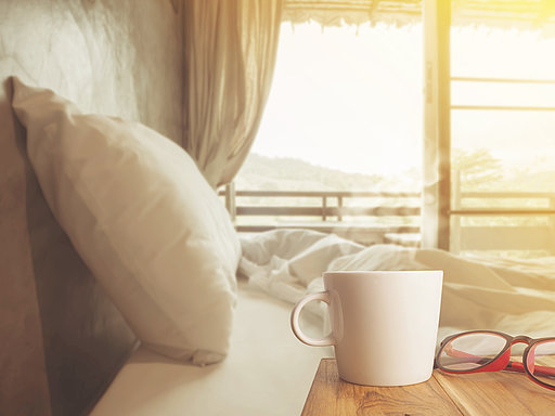 Leeres Bett im Morgenlicht mit Kaffeetasse und Brille im Vordergrund