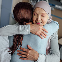 Jüngere Frau umarmt ältere Frau mit Kopftuch in einem Krankenhauszimmer. 