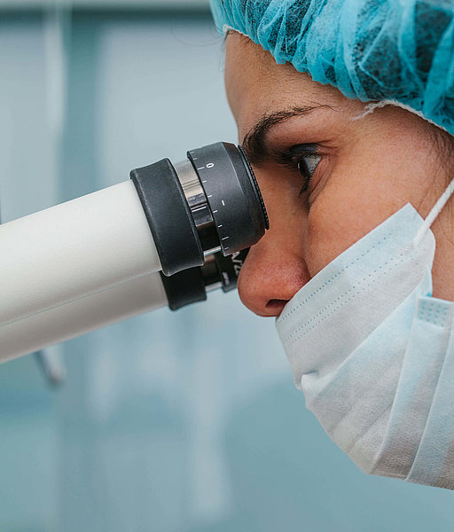 Frau mit Mundschutz und OP-Haube guckt durch ein Mikroskop