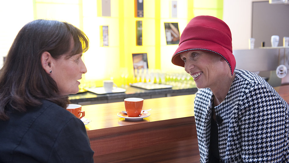 Brustkrebspatientin Angelika lächelt im Gespräch mit einer Bekannten beim Kaffeetrinken
