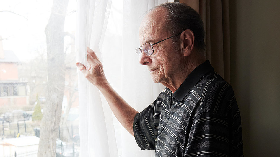 Ein älterer Mann zieht eine Gardine zur Seite und blickt aus dem Fenster