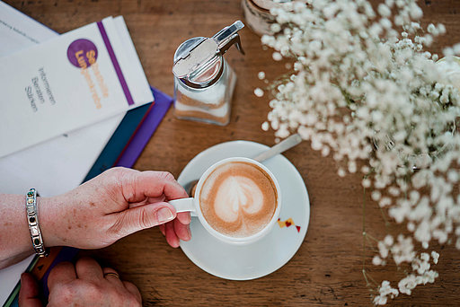 Ältere Hand hält eine Tasse mit Kaffee mit einem Blattmuster aus Milchschaum. 