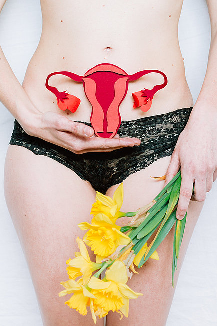 Eine Frau hält ein Modell weiblicher Geschlechtsorgane vor ihren Unterbauch und Blumen in der Hand