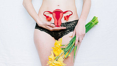 Eine Frau hält ein Modell weiblicher Geschlechtsorgane vor ihren Unterbauch und Blumen in der Hand