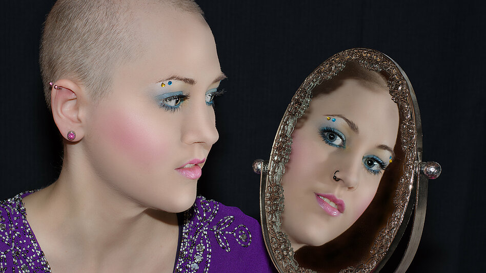 Krebs: junge Frau mit kurzen Haaren sieht in den Spiegel