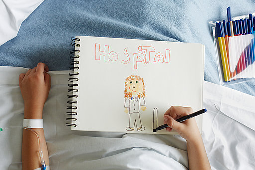 Ein Kind liegt in einem Klinikbett mit Malblock auf dem Schoß.