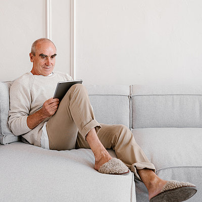 Ein älterer Mann entspannt auf dem Sofa mit einem Tabletcomputer in der Hand.