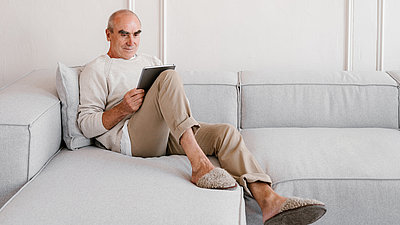 Ein älterer Mann entspannt auf dem Sofa mit einem Tabletcomputer in der Hand.