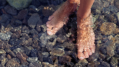 Füße und Knöchel stehen in klarem Wasser auf Kieselsteinen
