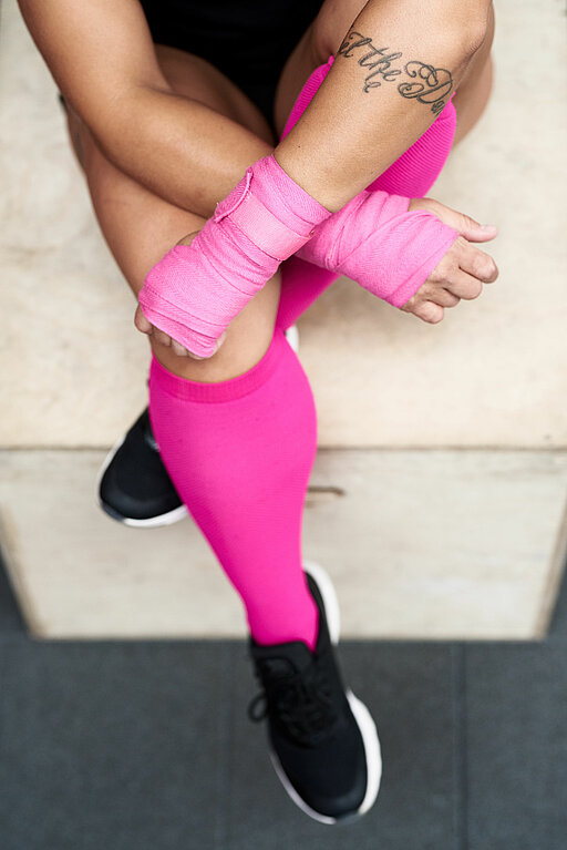 Ausschnitt einer sportlichen Person mit pinkfarbenen Bandagen um Beine und Hände.