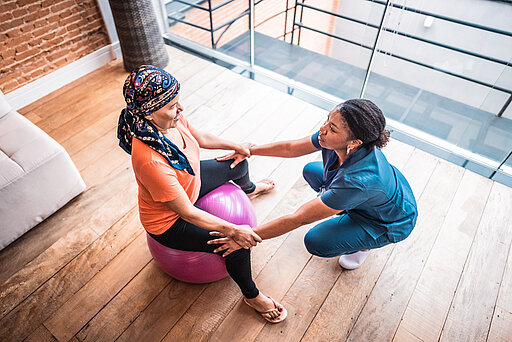 Eine erkrankte Frau sitzt auf einem Gummiball und macht zusammen mit einer Therapeutin eine Übung