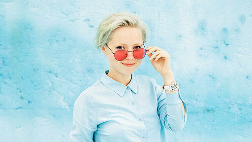 Frau in hellblauer Bluse steht vor einer hellblauen Wand und guckt durch eine Sonnenbrille mit roten Gläsern. 