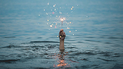 Aus einem Gewässer ragt eine Hand mit brennender Wunderkerze
