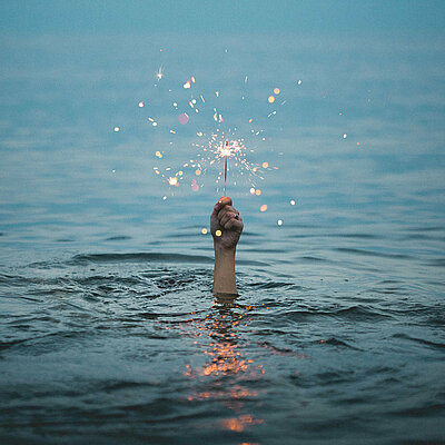 Aus einem Gewässer ragt eine Hand mit brennender Wunderkerze