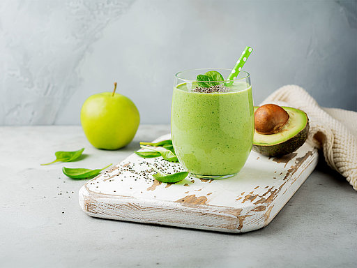 Ein grüner Smoothie im Glas neben einer halben Avocado, einem grünen Apfel und frischen Spinatblättern