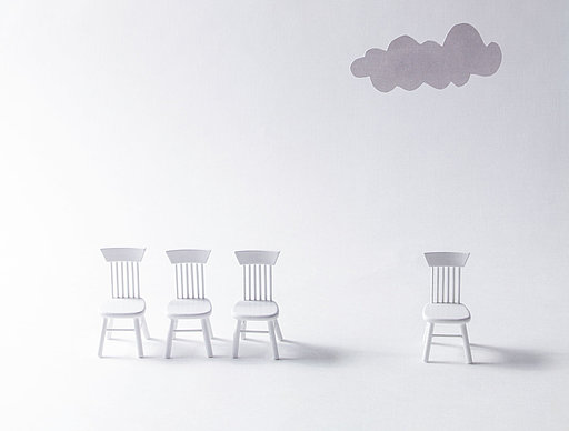 Vier Stühle nebeneinander aufgereiht, ein weiterer Stuhl steht abseits unter einer Gewitterwolke 