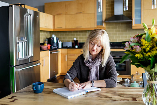 Frau sitzt am Küchentisch und schreibt in ein Notizbuch
