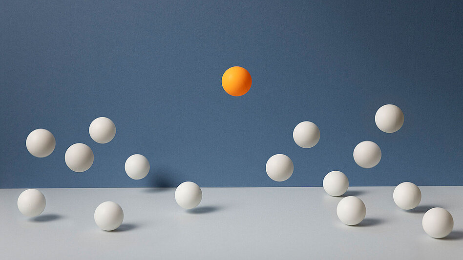 daskwort_metastasierter_brustkrebs_tumortestung Eine Gruppe von Tischtennisbällen springt auf blauem Hintergrund. Der orangefarbene Ball ist höher als die weißen Bälle.