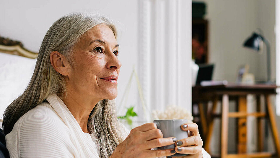 Eine ältere Frau mit einer Tasse in der Hand blickt lächelnd in die Ferne