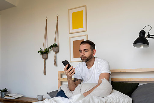 Mann mittleren Alters sitzt im Bett mit Handy in der Hand