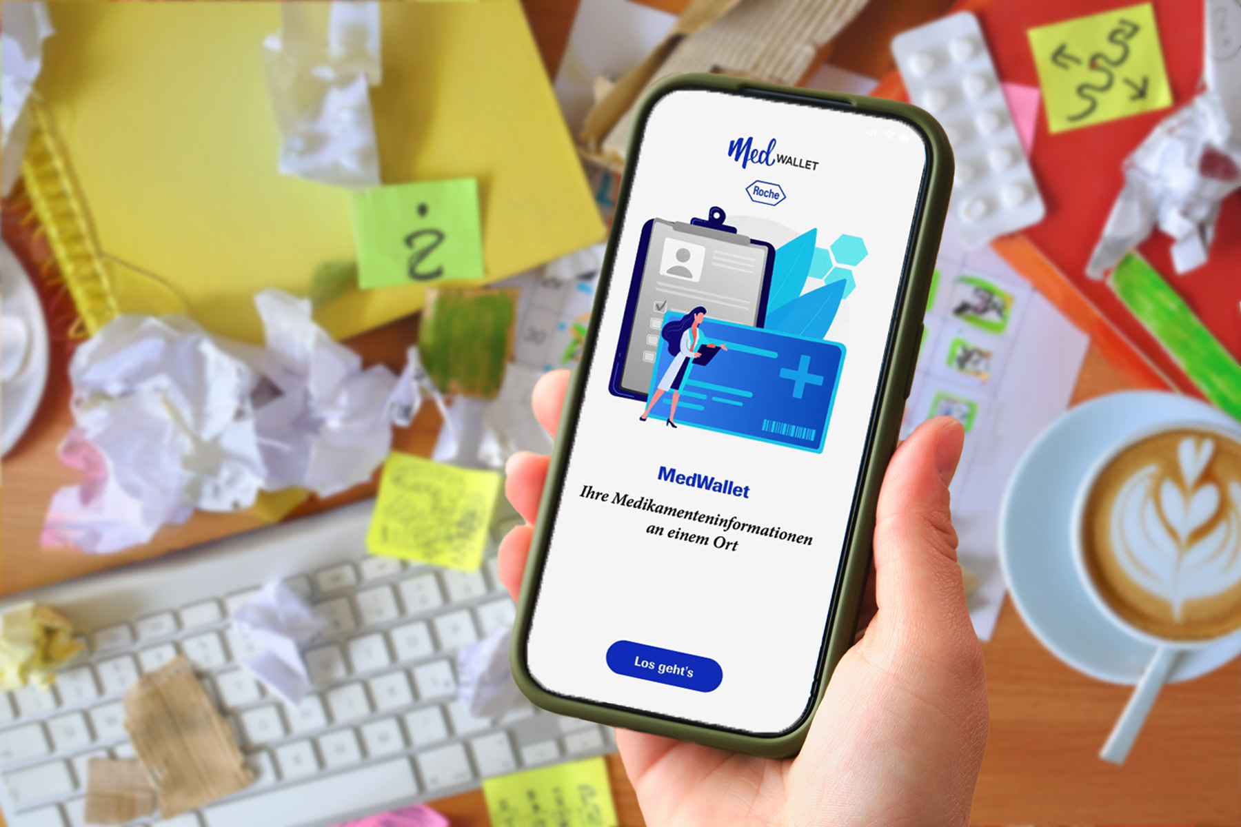 Person hält Smartphone mit geöffneter MedWallet-App; im Hintergrund unordentlicher Schreibtisch mit Notizzetteln, Tastatur und Kaffeetasse.